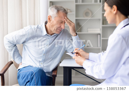 Senior male patient holding head in pain while female doctor writes notes 138053367