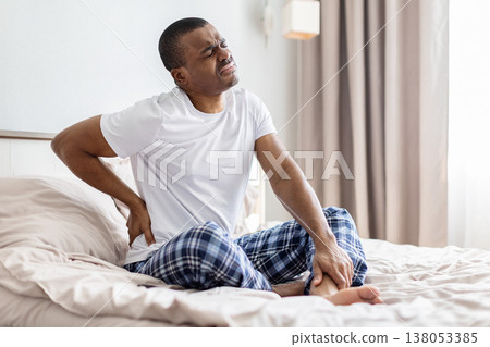 Young african american man sitting on bed holding lower back in pain Young african american man sitting on bed holding lower back in pain 138053385