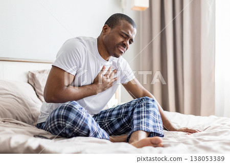 Young black man sitting on bed holding chest in pain, showing discomfort 138053389