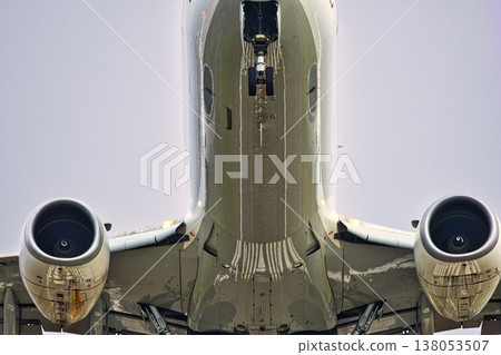 Detailed Underside View of Commercial Aircraft on Final Approach: Close-up of Jet Engines, Extended Nose Landing Gear, and Reflective Fuselage Surface against Overcast Sky Detailed Underside View of Commercial Aircraft on Final Approach: Close-up of Jet Engines, Extended Nose Landing Gear, and Reflective Fuselage Surface against Overcast Sky 138053507