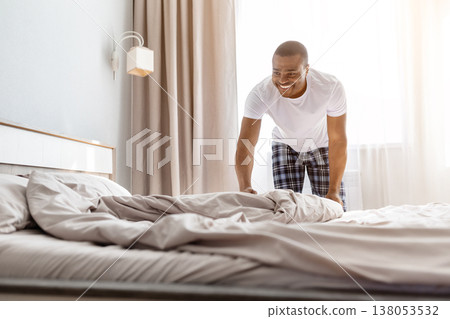 Young black man making bed in morning, arranging blanket and pillows Young black man making bed in morning, arranging blanket and pillows 138053532