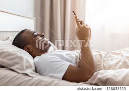 Young african american man lying in bed using smartphone and smiling Young african american man lying in bed using smartphone and smiling 138053533