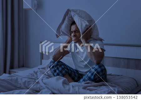 Young black man sitting in bed at night covering ears with pillow 138053542