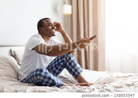 Young black man sitting on bed smiling while holding remote control Young black man sitting on bed smiling while holding remote control 138053547