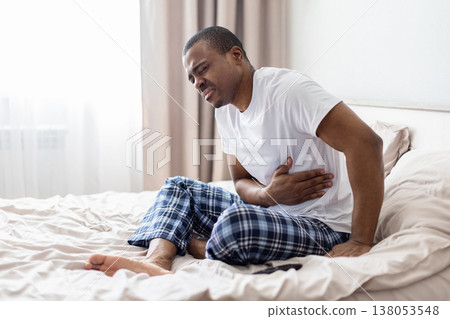 Young black man sitting on bed holding stomach with pain expression Young black man sitting on bed holding stomach with pain expression 138053548