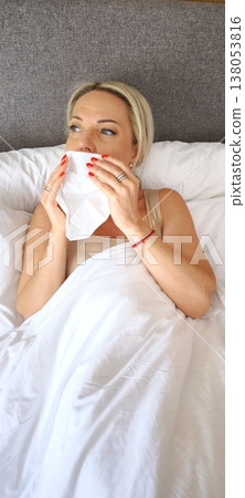 A woman blows her nose into a paper napkin while lying in bed A woman blows her nose into a paper napkin while lying in bed 138053816