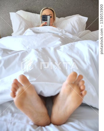 Cozy morning, woman lies in bed and looks at her phone, only her feet are visible 138053900