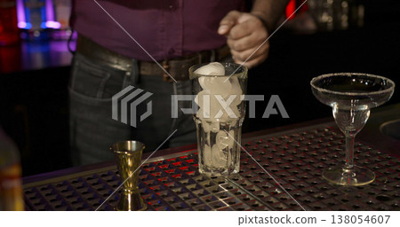 Bartender prepares cocktail with ice and glassware at bar counter during evening hours Bartender prepares cocktail with ice and glassware at bar counter during evening hours 138054607