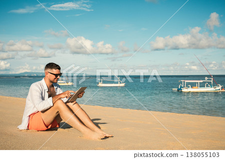 Man sits on the sand using a tablet near the water 138055103