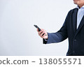 A man in a suit, whose face is obscured, is shown looking at a smartphone against a white background, photographed from an angle. 138055571