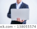 A blurred image of a faceless man in a suit standing in front of a white background, holding a laptop. 138055574