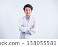 A male doctor in his 30s, wearing a white coat, stands with his arms crossed in front of a white background, looking directly at the camera. 138055581