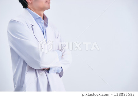 A faceless male doctor in his 30s, wearing a white coat and with his arms crossed, photographed from the side against a white background. 138055582
