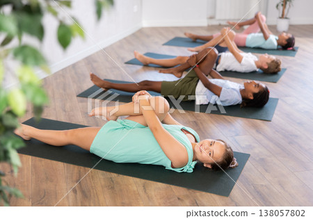 Children doing yoga with group in studio, lying on mat with knee pressed to chest with hands, performing hamstring and low back stretching Ardha Pawanmuktasana 138057802