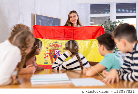 Smiling young female teacher explaining culture of country while showing Spanish flag to schoolchildren preteens in classroom Smiling young female teacher explaining culture of country while showing Spanish flag to schoolchildren preteens in classroom 138057821