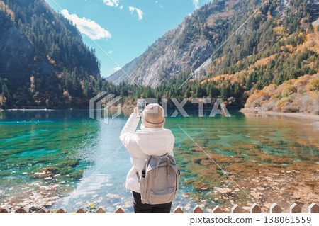 Happy woman traveler travel in jiuzhaigou national park in Sichuan, China. Tourist sightseeing and taking photo beautiful lake by mobile phone in Autumn season. Famous landmark near Chengdu, China Happy woman traveler travel in jiuzhaigou national park in Sichuan, China. Tourist sightseeing and taking photo beautiful lake by mobile phone in Autumn season. Famous landmark near Chengdu, China 138061559