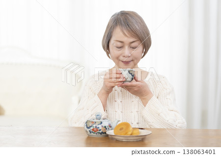A senior woman drinking tea and snacks in the living room. A senior woman drinking tea and snacks in the living room. 138063401