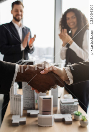 Diverse business professionals shaking hands over architectural models, with colleagues applauding in the background, signifying a successful partnership or project completion Diverse business professionals shaking hands over architectural models, with colleagues applauding in the background, signifying a successful partnership or project completion 138064105