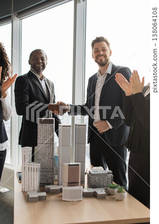 Diverse team celebrates a signed real estate agreement over an architectural scale model in a bright office setting to mark a successful deal and partnership for development projects 138064108