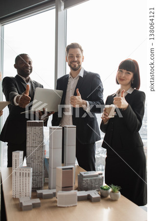 A diverse group of professionals in business attire gives a thumbs-up gesture while standing near architectural models and a laptop 138064121