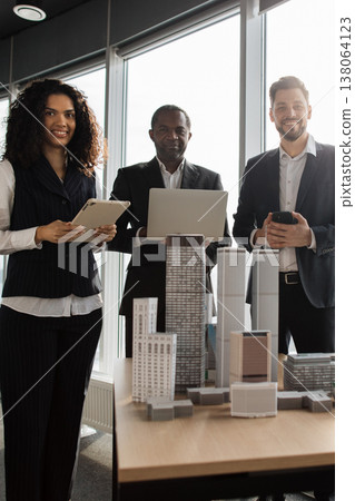 A professional and diverse team of three business people stand behind an architectural model, holding technology devices and looking towards the camera with smiles 138064123