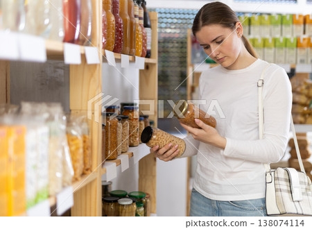 Girl customer in supermarket examines jar of kidney bean and lentil, chooses food-stuff in grocery Girl customer in supermarket examines jar of kidney bean and lentil, chooses food-stuff in grocery 138074114