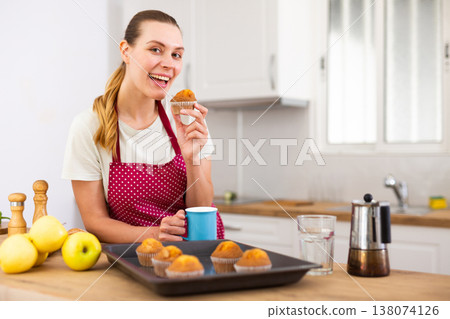 Smiling young woman in apron eating cupcakes from baking tray 138074126