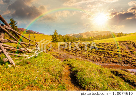 sunset over mountainous countryside landscape in spring. wooden fence and grassy meadow on a green hill. distant mountain ridge with snow capped tops. cloudy sky and rainbow 138074610