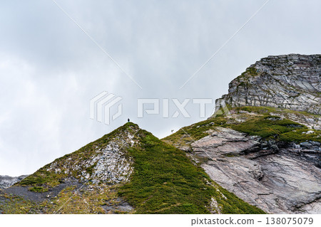 Man on top of a mountain with rocky peaks and green grass under a vast gray sky, exploring adventure and overcoming challenges, copy space Man on top of a mountain with rocky peaks and green grass under a vast gray sky, exploring adventure and overcoming challenges, copy space 138075079