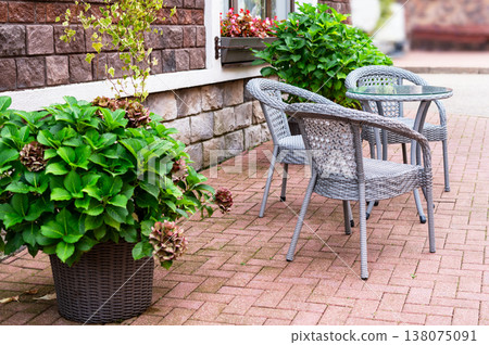 Outdoor cafe patio with wicker furniture set including table and chair on stone paved ground. Cozy restaurant terrace with green plant in pot near brick wall for exterior design concept. Outdoor cafe patio with wicker furniture set including table and chair on stone paved ground. Cozy restaurant terrace with green plant in pot near brick wall for exterior design concept. 138075091
