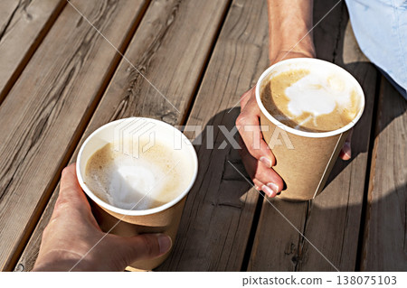 Hands of two people holding beige paper cups of cappuccino on a wooden striped table outside, drinking coffee, meeting friends 138075103