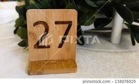close up of a wooden stand sign with the number 27 written on it. Plants at the back on a white table with copy space close up of a wooden stand sign with the number 27 written on it. Plants at the back on a white table with copy space 138076020