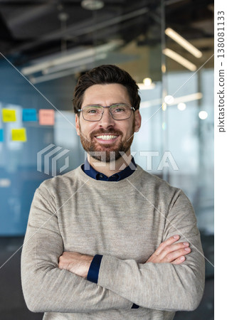 Confident businessman wearing glasses and a beard smiling at the camera, portraying professionalism, success, and positive leadership in a modern office environment 138081133