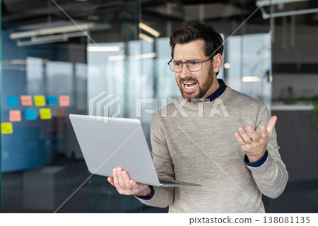 Businessman experiencing stress and disappointment from a computer error, screaming at his laptop screen in an office environment, symbolizing modern work challenges 138081135
