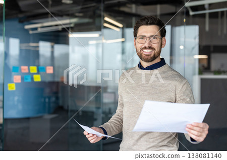 Young businessman wearing eyeglasses and a sweater, smiling confidently while holding papers with a bright professional office interior as the background Young businessman wearing eyeglasses and a sweater, smiling confidently while holding papers with a bright professional office interior as the background 138081140