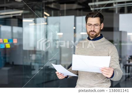 Young businessman standing in a contemporary glass office, reading and analyzing financial documents with a focused expression, representing data review and corporate management Young businessman standing in a contemporary glass office, reading and analyzing financial documents with a focused expression, representing data review and corporate management 138081141