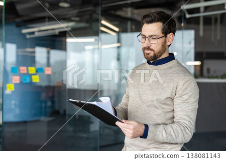 Professional man wearing glasses and a beard, focusing intently while reviewing documents on a clipboard, standing in a modern office with glass walls and colorful sticky notes Professional man wearing glasses and a beard, focusing intently while reviewing documents on a clipboard, standing in a modern office with glass walls and colorful sticky notes 138081143