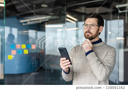 Thoughtful businessman with a beard and glasses holding a smartphone, looking away in contemplation, working on new business strategies and ideas in a modern office Thoughtful businessman with a beard and glasses holding a smartphone, looking away in contemplation, working on new business strategies and ideas in a modern office 138081162