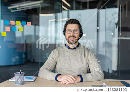 Smiling male call center agent wearing a headset and glasses, sitting at his desk in a modern office, providing distant customer service and contact support Smiling male call center agent wearing a headset and glasses, sitting at his desk in a modern office, providing distant customer service and contact support 138081193