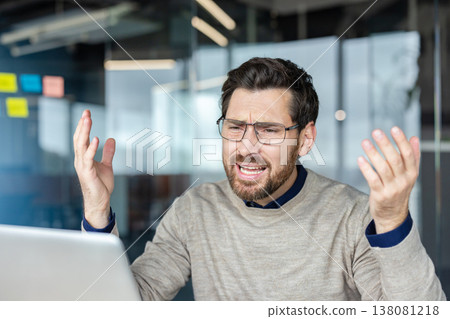 Young businessman clutching his head in frustration at a laptop on a cluttered desk, showing stress and overwhelm during a difficult work deadline in a modern office setting Young businessman clutching his head in frustration at a laptop on a cluttered desk, showing stress and overwhelm during a difficult work deadline in a modern office setting 138081218