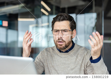 Man with glasses, experiencing frustration and stress while working on a laptop, raising his hands in a gesture of confusion and anger in a modern office setting 138081219