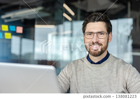 Smiling professional business man wearing glasses and a beard, working confidently on a laptop in a modern office, representing successful career and technology Smiling professional business man wearing glasses and a beard, working confidently on a laptop in a modern office, representing successful career and technology 138081223