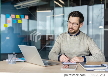 Businessman wearing glasses diligently working on a laptop computer and taking notes, in a notebook at his desk in a contemporary co-working office space with glass walls Businessman wearing glasses diligently working on a laptop computer and taking notes, in a notebook at his desk in a contemporary co-working office space with glass walls 138081231