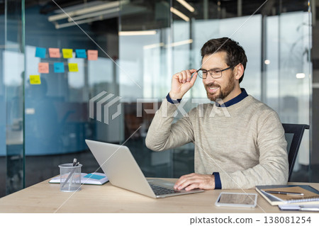 Happy professional businessman in smart casual clothes adjusting glasses while engaging with a laptop, managing tasks and smiling in a contemporary workspace with a glass wall and sticky notes Happy professional businessman in smart casual clothes adjusting glasses while engaging with a laptop, managing tasks and smiling in a contemporary workspace with a glass wall and sticky notes 138081254