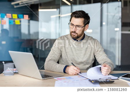 Professional man wearing glasses working on a laptop at his desk in a modern office, concentrating while reviewing financial documents, project schedules, and data analytics paperwork 138081260