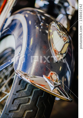 A vertical close-up of a blue metallic motorcycle fender featuring a classic chrome "eagle" ornament, a vintage light, and a tire with a white sidewall. A vertical close-up of a blue metallic motorcycle fender featuring a classic chrome "eagle" ornament, a vintage light, and a tire with a white sidewall. 138081542