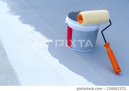 Orange paint roller resting on a bucket of grey liquid coating on a rooftop or floor. A white stripe of primer is visible on the textured surface. Concept of waterproofing and renovation. Orange paint roller resting on a bucket of grey liquid coating on a rooftop or floor. A white stripe of primer is visible on the textured surface. Concept of waterproofing and renovation. 138081572