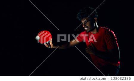 Ping pong player striking ball with powerful motion in dark dramatic lighting. Ping pong player striking ball with powerful motion in dark dramatic lighting. 138082386
