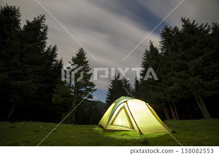 Summer camping at night. Illuminated tourist tent on green clearing on distant mountain background. Summer camping at night. Illuminated tourist tent on green clearing on distant mountain background. 138082553