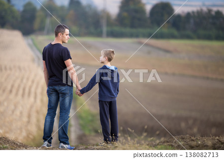 Profile portrait of young father and smiling son standing in grassy field holding hands on blurred green trees and blue sky background. Active lifestyle, family relations, weekend activity concept. Profile portrait of young father and smiling son standing in grassy field holding hands on blurred green trees and blue sky background. Active lifestyle, family relations, weekend activity concept. 138082713